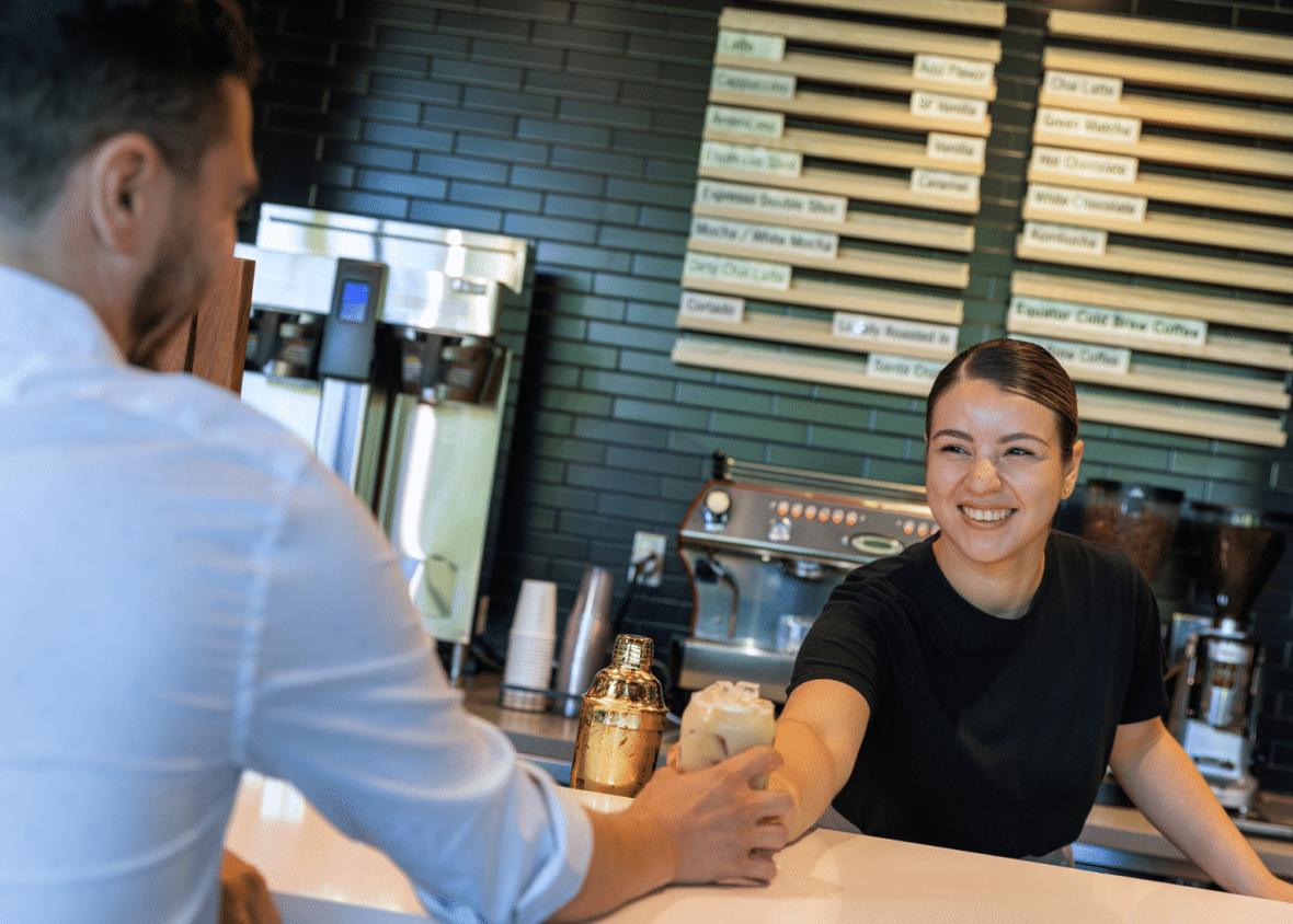 Female barista smiling and sliding iced coffee across the counter to a customer