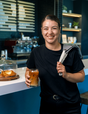 Female barista smiling and holding coffee with an apron thrown over her shoulder